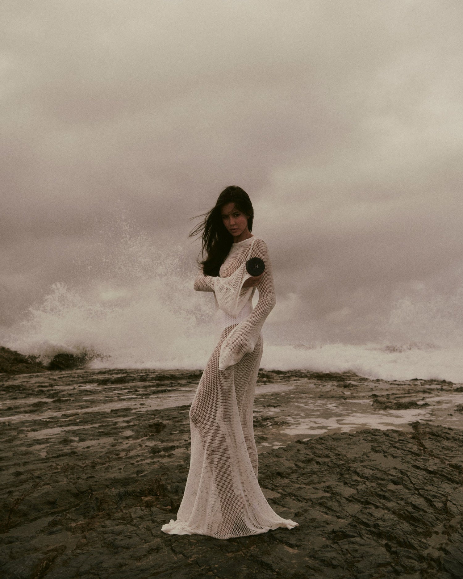 Woman in a white dress standing on a rocky beach with waves crashing behind her under a stormy sky.